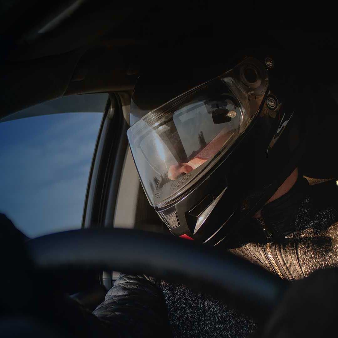 A race car driver grips the wheel, clad in a black helmet with a gleaming face shield.